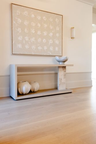 Entryway featuring light oak hardwood flooring, a minimalist beige console table with sculptural vases, and abstract art with raised white floral motifs.
