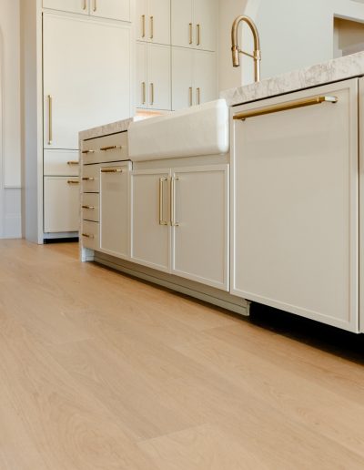 Kitchen with light oak flooring, white shaker cabinets, gold handles, and a farmhouse sink beneath marble countertops.