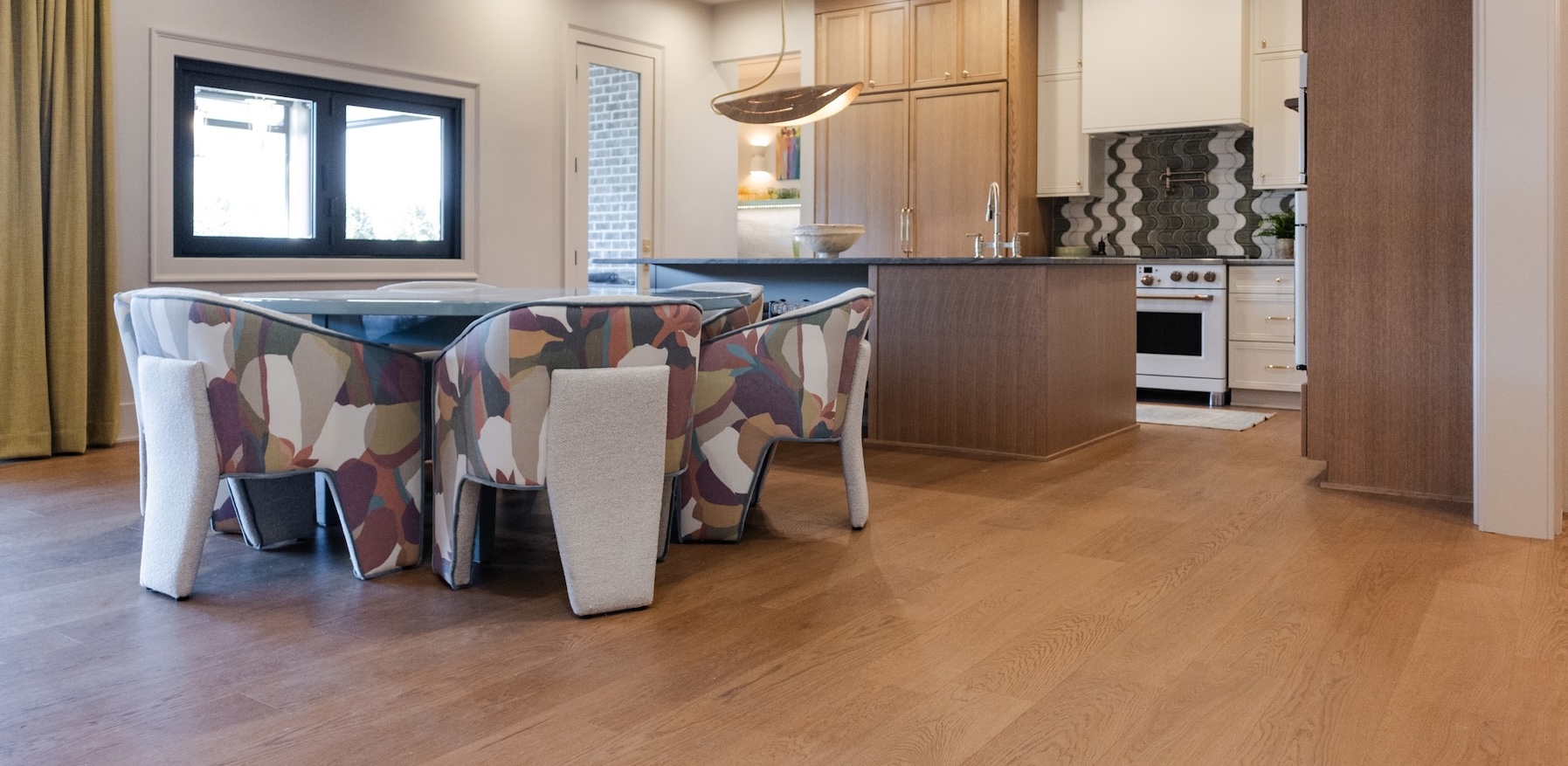 European white oak flooring flows through an open dining room and kitchen featuring colorful upholstered chairs and warm natural wood cabinetry.