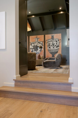 Warm European white oak flooring leads into a moody lounge with dark walls, exposed ceiling beams, and sculptural seating.